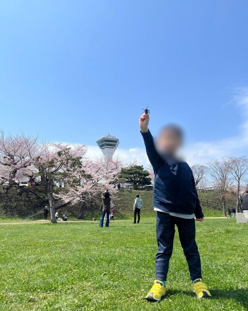 トミカのヘリコプターのおもちゃを空にかざす子ども、背景に桜満開の五稜郭タワーと堀沿いの桜並木