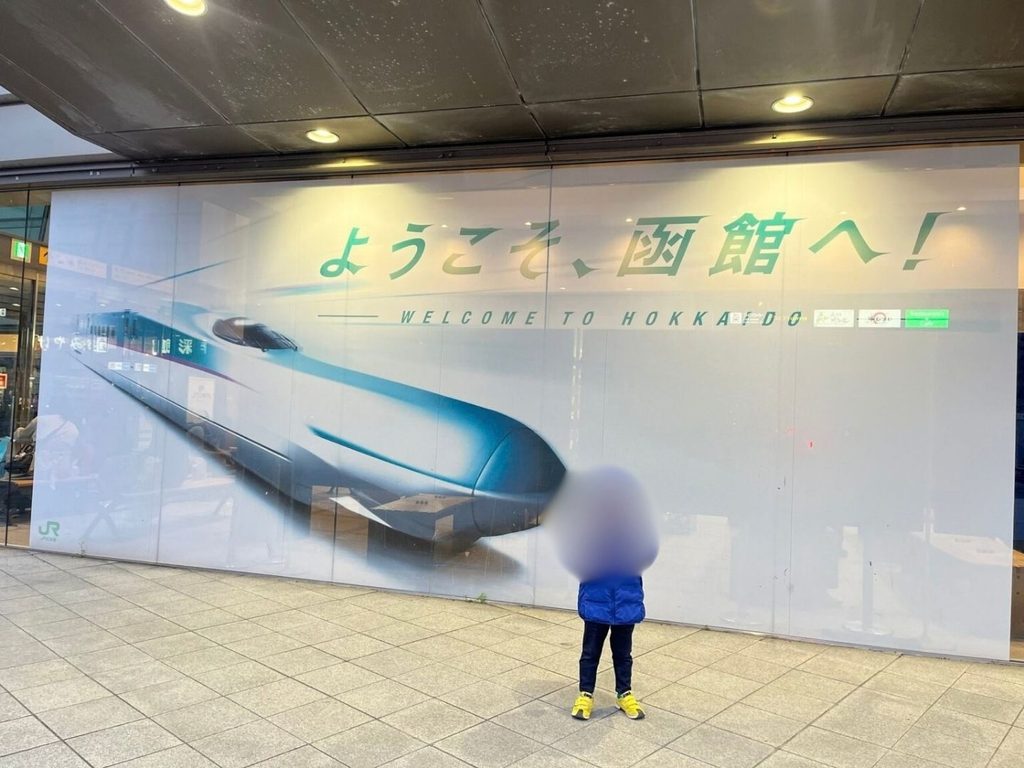 「ようこそ、函館へ!」の新幹線ウェルカムボードの前に立つ子ども